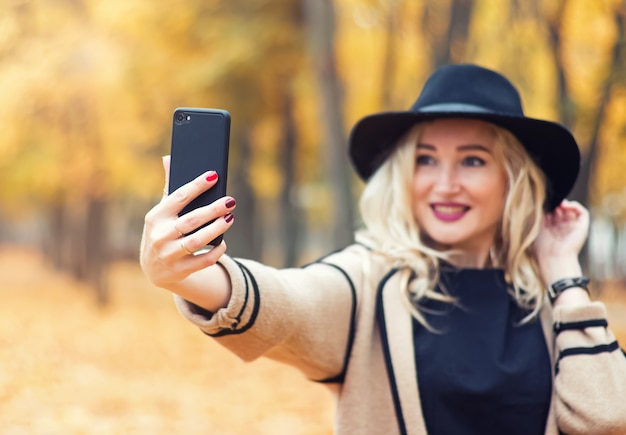 Mujer bonita joven con sombrero negro haciendo selfie con su teléfono inteligente en el parque de otoño