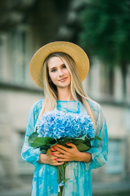 Mujer bonita joven con sombrero de paja está sonriendo con hortensias en la calle de verano