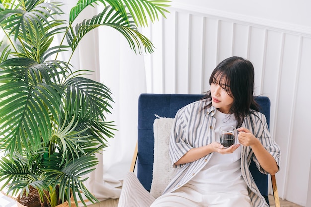 Mujer bonita joven sonríe y se sienta en la silla de la sala de estar moderna con una planta verde Relajándose tomando una taza de café caliente y disfrutando de un momento tranquilo y un ambiente interior en un hogar acogedor