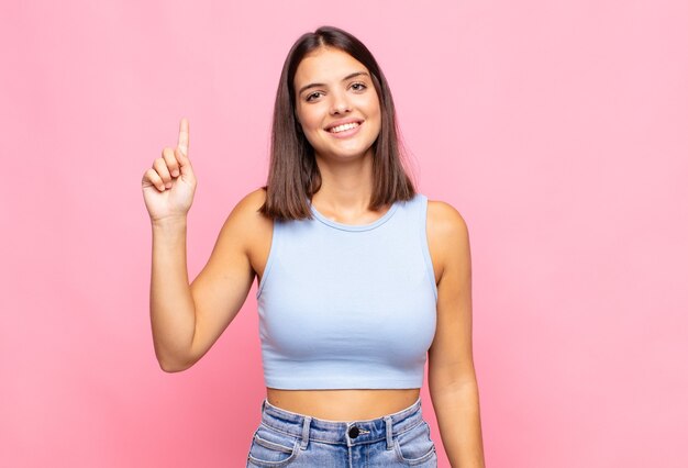 Mujer bonita joven sonriendo alegre y felizmente, apuntando hacia arriba con una mano para copiar el espacio