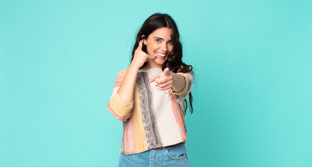 Mujer bonita joven sonriendo alegremente y apuntando a la cámara mientras hace una llamada, gesto más tarde, hablando por teléfono