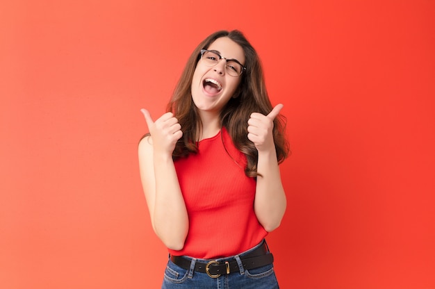 Foto mujer bonita joven sonriendo alegremente y luciendo feliz, sintiéndose despreocupada y positiva con ambos pulgares contra la pared roja