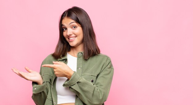 Mujer bonita joven sonriendo alegremente y señalando para copiar el espacio en la palma en el lateral, mostrando o publicitando un objeto