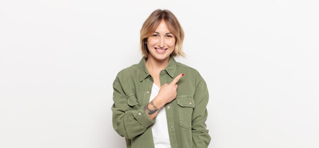 Mujer bonita joven sonriendo alegremente, sintiéndose feliz y apuntando hacia un lado y hacia arriba, mostrando el objeto en el espacio de la copia