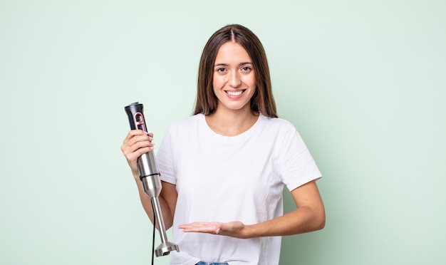 Mujer bonita joven sonriendo alegremente, sintiéndose feliz y mostrando un concepto. concepto de batidora de mano