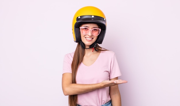 Mujer bonita joven sonriendo alegremente, sintiéndose feliz y mostrando un concepto. motociclista y casco