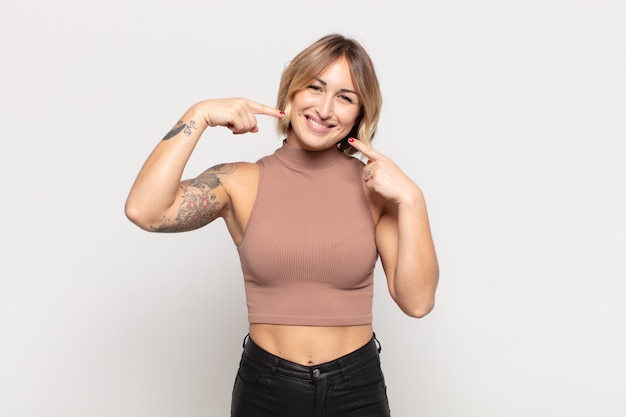 Mujer bonita joven sonriendo con confianza apuntando a su propia sonrisa amplia, actitud positiva, relajada y satisfecha