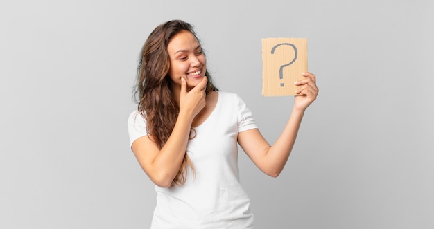 Mujer bonita joven sonriendo con una expresión feliz y segura con la mano en la barbilla y sosteniendo un signo de interrogación