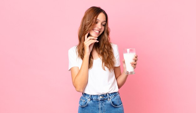 Mujer bonita joven sonriendo con una expresión feliz y segura con la mano en la barbilla y sosteniendo un vaso de leche