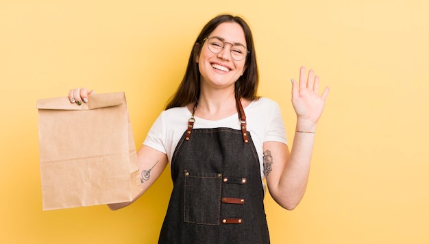 Mujer bonita joven sonriendo felizmente agitando la mano dándole la bienvenida y saludándolo mensajero de comida rápida