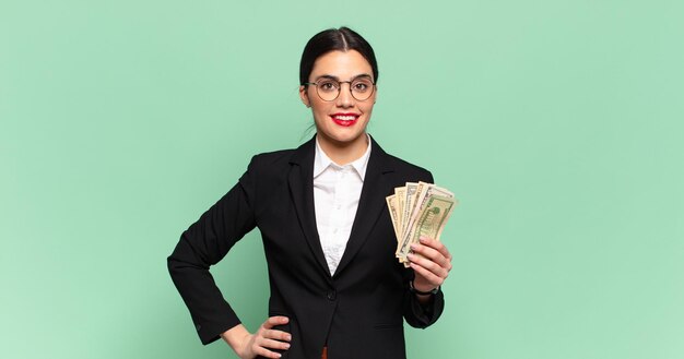 Mujer bonita joven sonriendo felizmente con una mano en la cadera y actitud confiada, positiva, orgullosa y amistosa. concepto de negocio y billetes