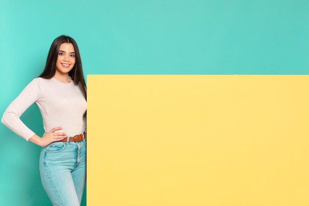 Foto mujer bonita joven sonriendo felizmente con una mano en la cadera y actitud confiada, positiva, orgullosa y amistosa. copia espacio para colocar tu concepto