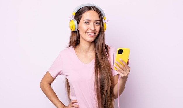 Mujer bonita joven sonriendo felizmente con una mano en la cadera y confiada. auriculares y smartphone