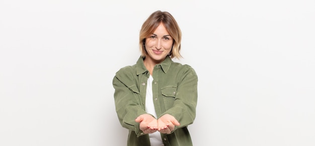 Mujer bonita joven sonriendo felizmente con mirada amistosa, segura y positiva, ofreciendo y mostrando un objeto o concepto