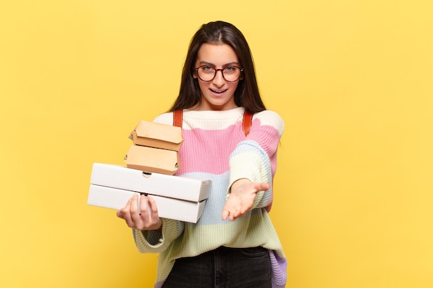 Mujer bonita joven sonriendo felizmente con mirada amistosa, segura y positiva, ofreciendo y mostrando un objeto o concepto