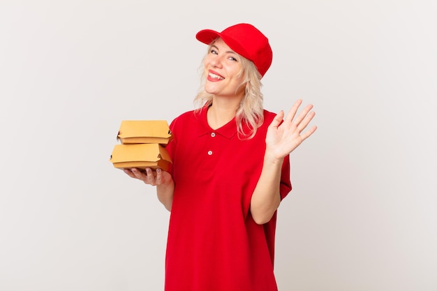 Mujer bonita joven sonriendo felizmente, saludando con la mano, dándote la bienvenida y saludándote. concepto de entrega de hamburguesas