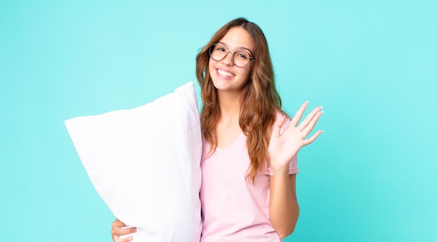 Mujer bonita joven sonriendo felizmente, saludando con la mano, dándote la bienvenida y saludándote en pijama y sosteniendo una almohada