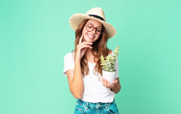 Mujer bonita joven sonriendo felizmente y soñando despierta o dudando con un sombrero de paja y sosteniendo un cactus