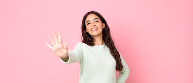 Mujer bonita joven sonriendo y mirando amigable, mostrando el número cinco o quinto con la mano hacia adelante, contando hacia atrás