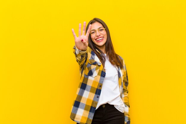 Mujer bonita joven sonriendo y mirando amigable, mostrando el número cuatro o cuarto con la mano hacia adelante, contando contra la pared naranja