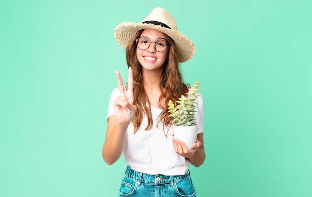 Mujer bonita joven sonriendo y mirando amigable, mostrando el número dos con un sombrero de paja y sosteniendo un cactus