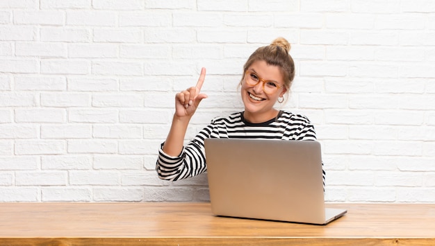 Mujer bonita joven sonriendo y mirando amigable, mostrando el número uno o primero con la mano hacia adelante, contando sentado con su computadora portátil