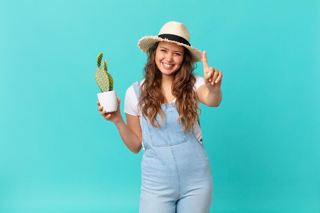 Mujer bonita joven sonriendo y mirando amigable, mostrando el número uno y sosteniendo un cactus