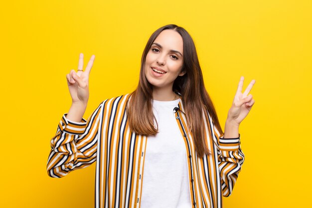 Mujer bonita joven sonriendo y mirando feliz, amigable y satisfecha, gesticulando victoria o paz con ambas manos contra la pared naranja