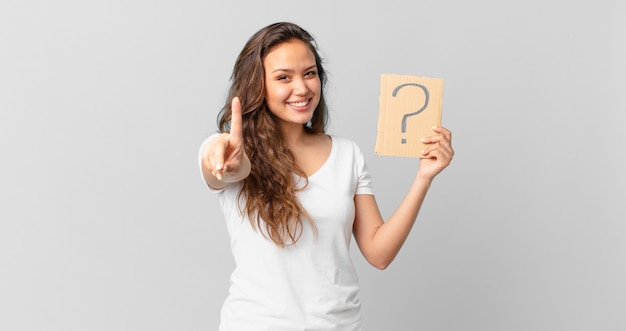 Mujer bonita joven sonriendo con orgullo y confianza haciendo el número uno y sosteniendo un signo de interrogación