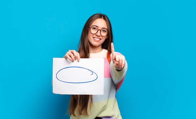 Mujer bonita joven sonriendo con orgullo y confianza haciendo la pose número uno triunfalmente, sintiéndose como un líder