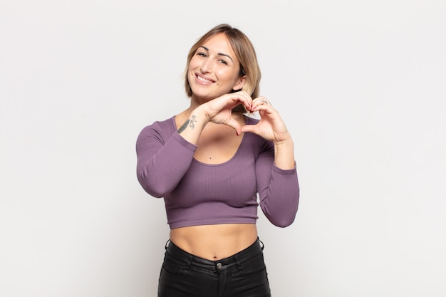 Mujer bonita joven sonriendo y sintiéndose feliz, linda, romántica y enamorada, haciendo forma de corazón con ambas manos