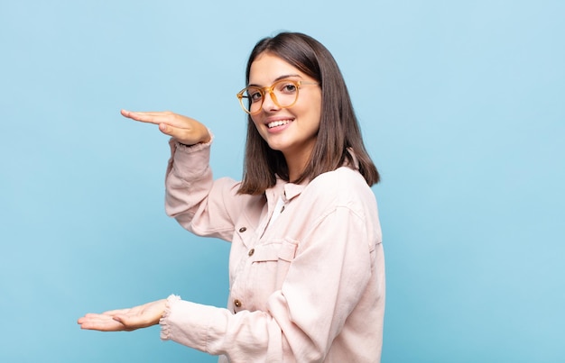 Mujer bonita joven sonriendo, sintiéndose feliz, positiva y satisfecha, sosteniendo o mostrando un objeto o concepto en el espacio de la copia