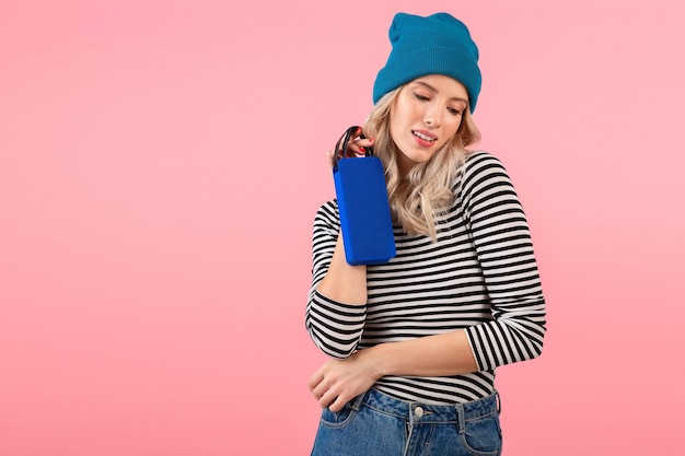 Mujer bonita joven sosteniendo un altavoz inalámbrico escuchando música con camisa a rayas y sombrero azul sonriendo feliz estado de ánimo positivo posando sobre fondo rosa aislado