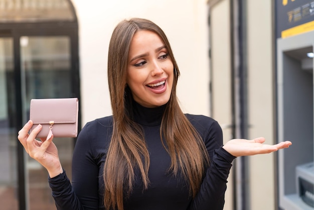Mujer bonita joven sosteniendo una billetera al aire libre con expresión facial sorpresa