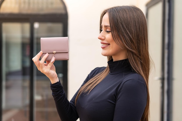 Mujer bonita joven sosteniendo una billetera al aire libre con expresión feliz