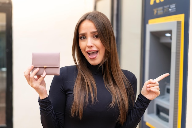 Mujer bonita joven sosteniendo una billetera al aire libre sorprendida y señalando con el dedo hacia un lado