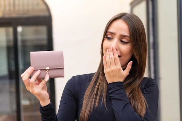 Mujer bonita joven sosteniendo una billetera al aire libre con sorpresa y expresión facial conmocionada