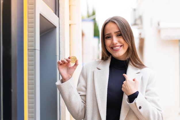 Mujer bonita joven sosteniendo un Bitcoin al aire libre con expresión facial sorpresa