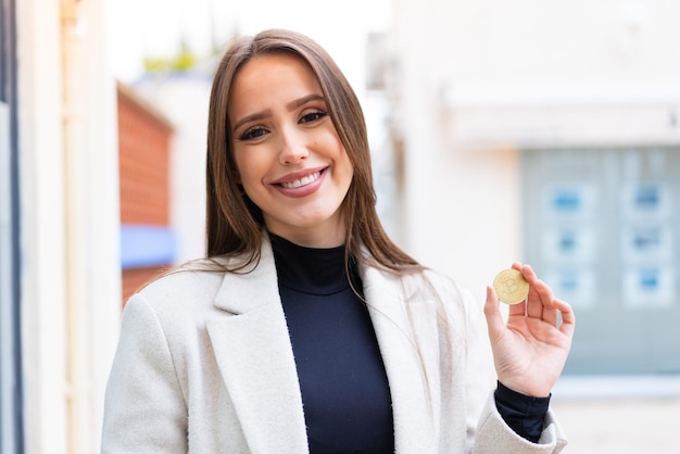 Mujer bonita joven sosteniendo un Bitcoin al aire libre sonriendo mucho