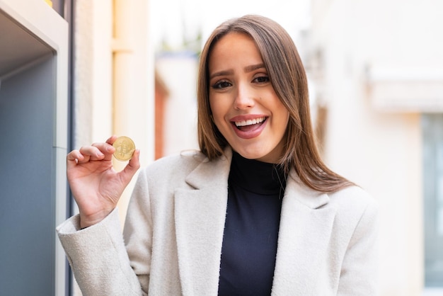 Mujer bonita joven sosteniendo un Bitcoin al aire libre con sorpresa y expresión facial conmocionada