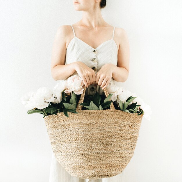 Mujer bonita joven sosteniendo una bolsa de paja con flores de peonía blanca sobre superficie blanca