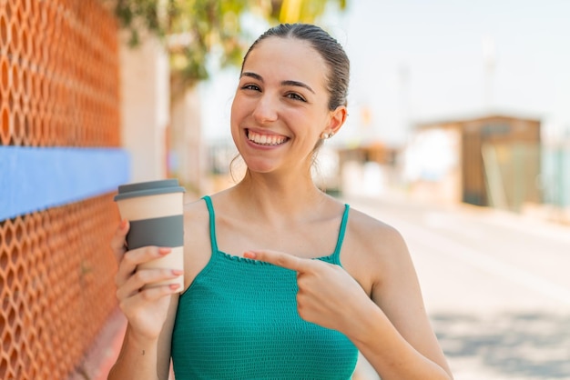 Mujer bonita joven sosteniendo un café para llevar al aire libre y apuntándolo