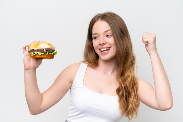 Mujer bonita joven sosteniendo una hamburguesa aislada sobre fondo blanco celebrando una victoria