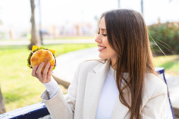 Mujer bonita joven sosteniendo una hamburguesa al aire libre con expresión feliz