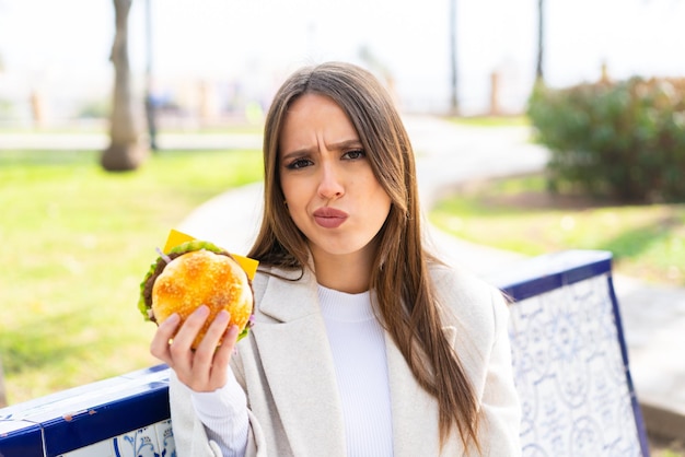 Mujer bonita joven sosteniendo una hamburguesa al aire libre con expresión triste