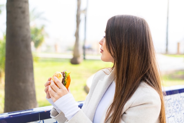 Mujer bonita joven sosteniendo una hamburguesa al aire libre en posición trasera