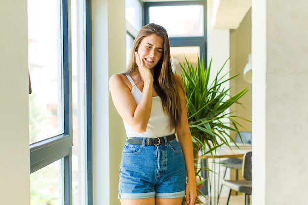 Mujer bonita joven sosteniendo la mejilla y sufriendo dolor de muelas, sintiéndose enferma, miserable e infeliz, en busca de un dentista