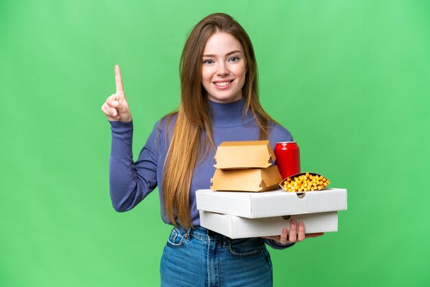 Mujer bonita joven sosteniendo pizzas y hamburguesas sobre un fondo de clave de croma aislado que muestra y levanta un dedo en señal de lo mejor