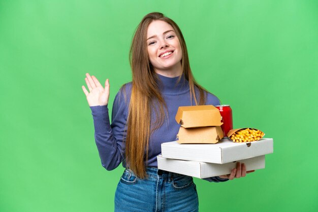 Mujer bonita joven sosteniendo pizzas y hamburguesas sobre un fondo clave de croma aislado saludando con la mano con expresión feliz