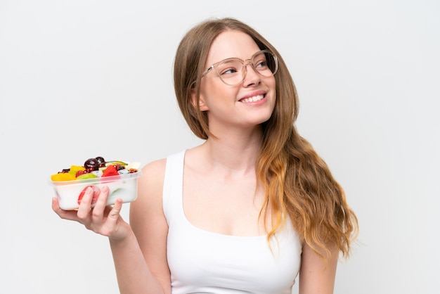 Mujer bonita joven sosteniendo un plato de fruta aislado sobre fondo blanco mirando hacia arriba mientras sonríe